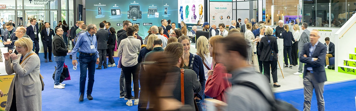 Viele Menschen laufen auf blauem Teppich durch eine große Halle mit Ständen von Industrieausstellern.