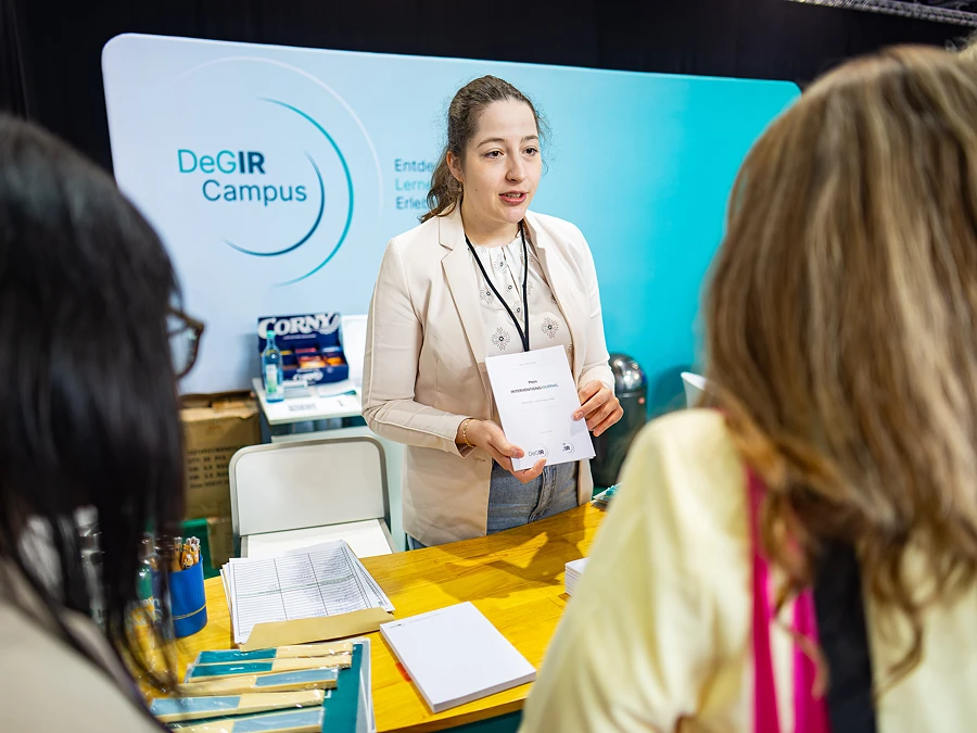 Eine junge Frau in einem hellen Blazer steht an einem Messestand des DeGIR Campus und hält ein Informationsblatt in der Hand, während sie mit Besuchern spricht.